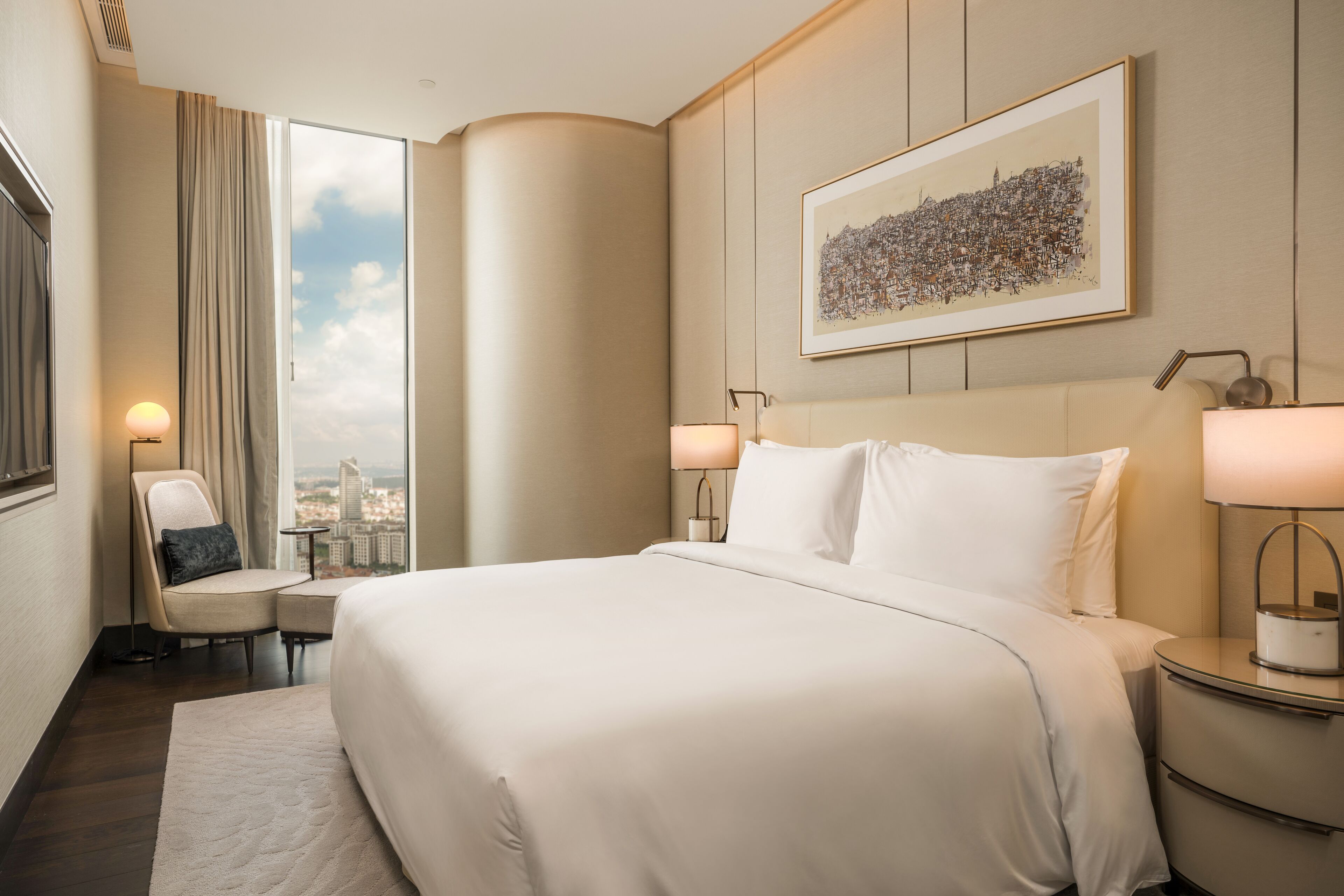 A hotel room with a large bed, a chair, a small table, and a nightstand. There is a window with a view of buildings and a framed cityscape on the wall.