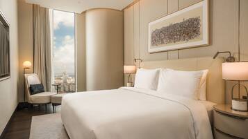 A hotel room with a large bed, a chair, a small table, and a nightstand. There is a window with a view of buildings and a framed cityscape on the wall.