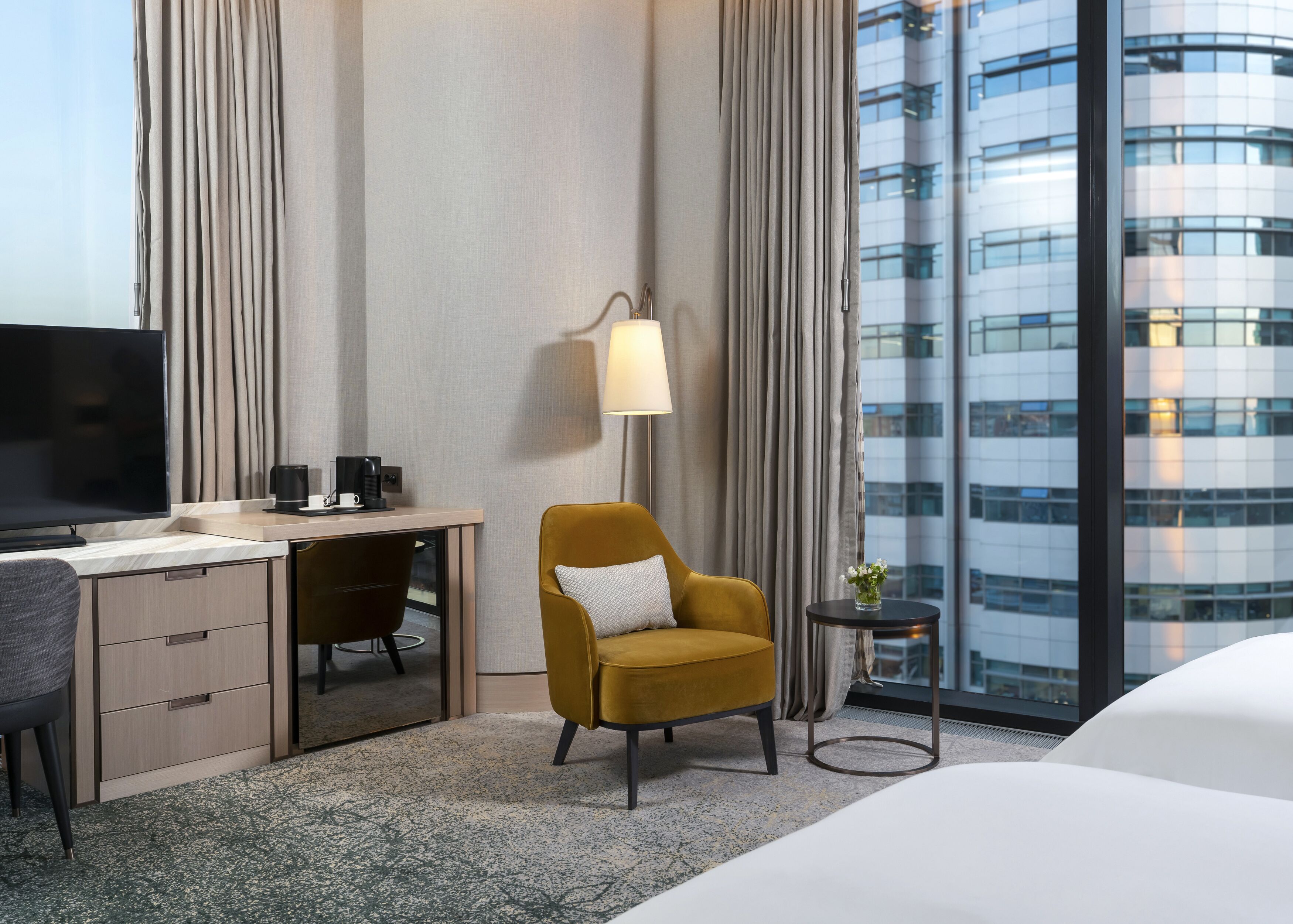 Een hotelkamer met een bed, een bureau met een televisie, een stoel, een lamp en uitzicht op een modern gebouw.