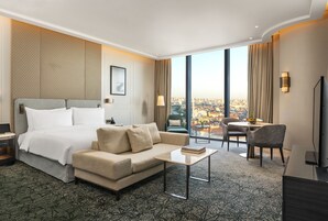 Habitación de hotel con cama, sofá, mesa de centro y zona de comedor con vistas a la ciudad.