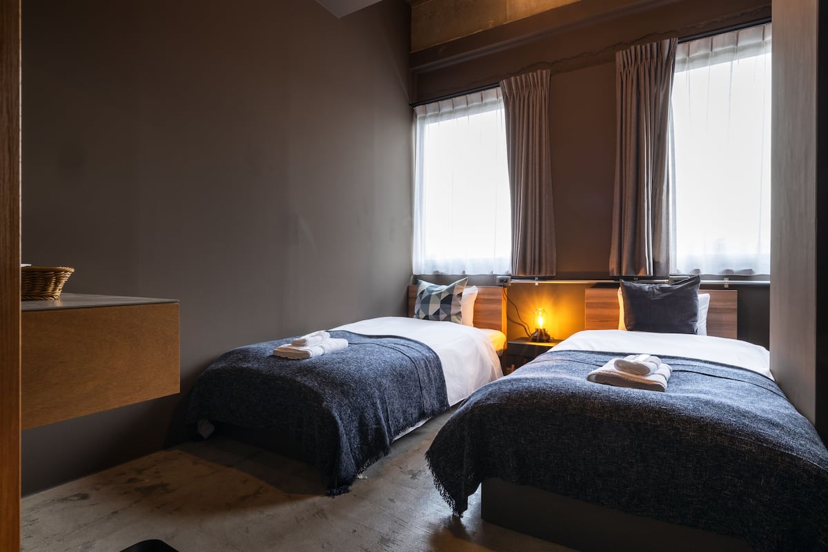 Habitación de hotel con dos camas, cabecero de madera, mesita de noche con lámpara y una ventana grande con cortinas.