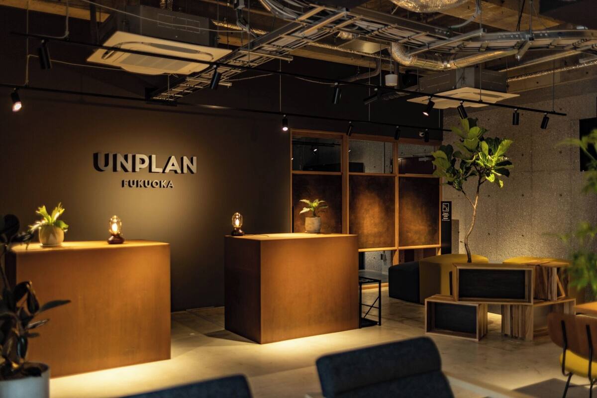 Un área de recepción con un mostrador de madera, plantas en macetas y un cartel que dice 'UNPLAN FUKUOKA'.