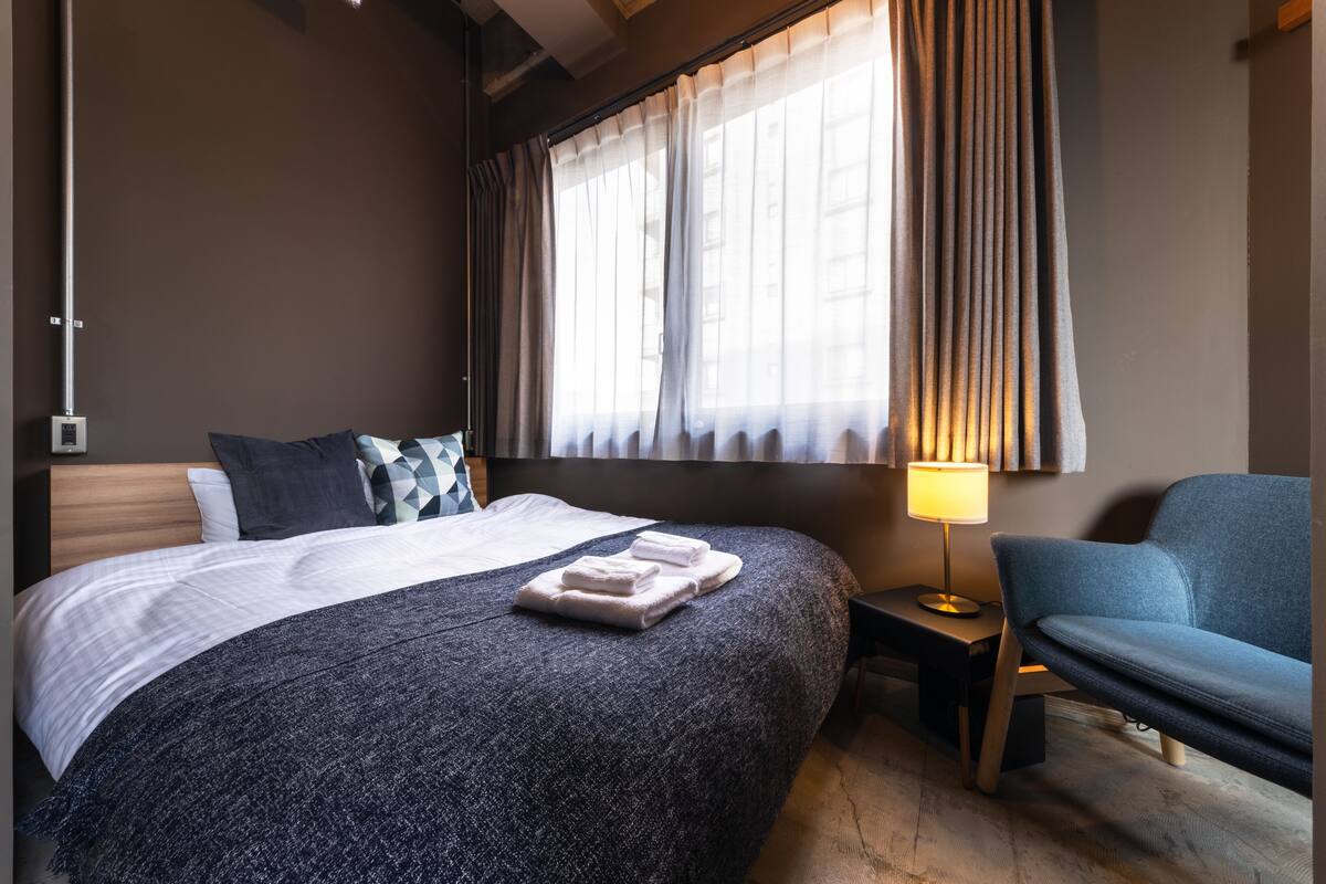 Una habitación de hotel con una cama grande, una mesita de noche con lámpara, una silla y una ventana con cortinas.