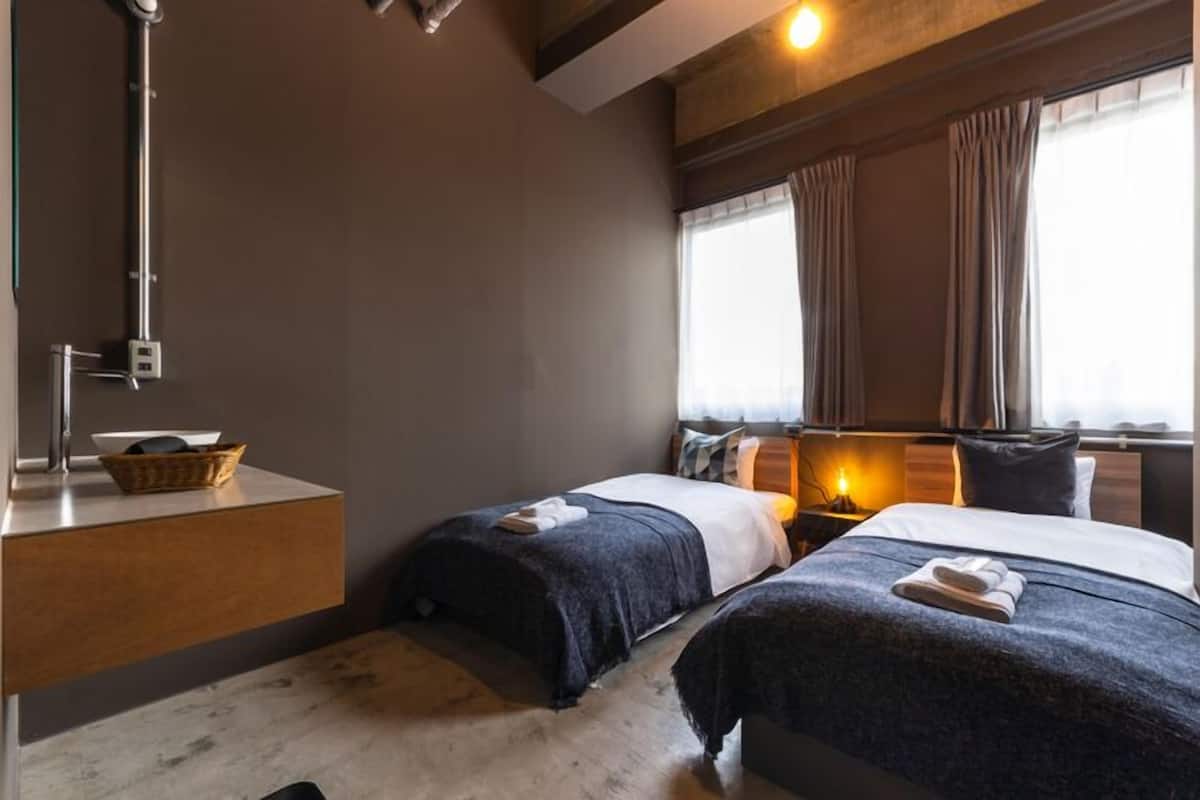 Habitación de hotel con dos camas, una mesita de noche, una lámpara, una ventana con cortinas y un perchero en la pared.
