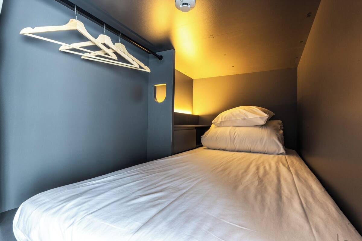 Una cama individual con ropa de cama blanca, un aplique en la pared y un juego de perchas de madera.