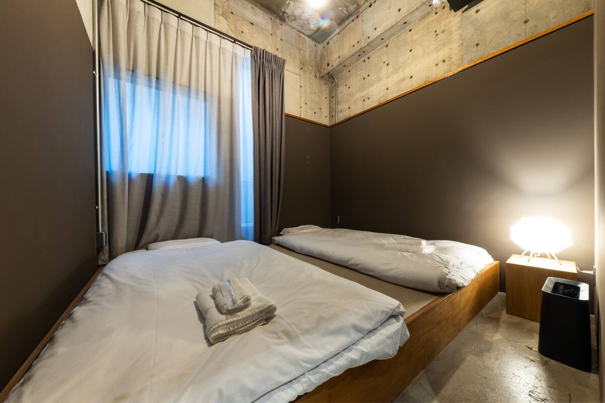 Habitación de hotel con dos camas, cabecero de madera, mesita de noche con lámpara y ventana con cortinas.
