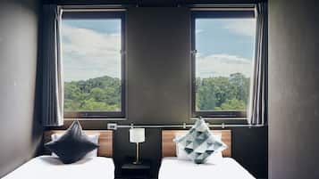 Habitación de hotel con dos camas, una mesita de noche con lámpara y vistas a un bosque a través de amplios ventanales.