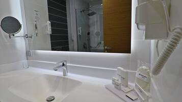 A modern bathroom with a white sink, a round mirror, a handheld showerhead, and a hairdryer mounted on the wall.