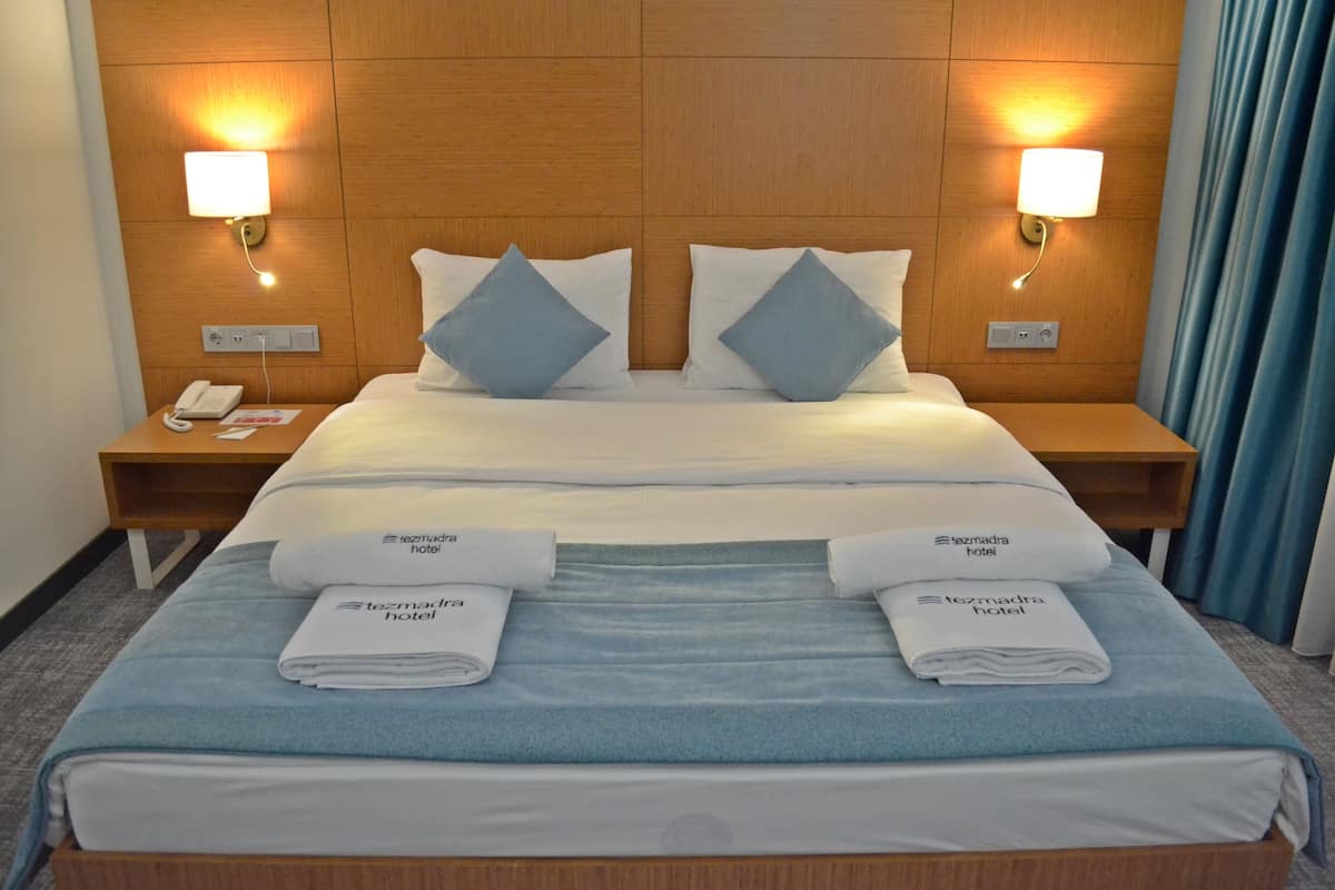 Una cama bien hecha con ropa de cama azul y blanca, dos almohadas y dos toallas dobladas con el nombre del hotel. Hay dos mesitas de noche, una con lámparas y un teléfono.