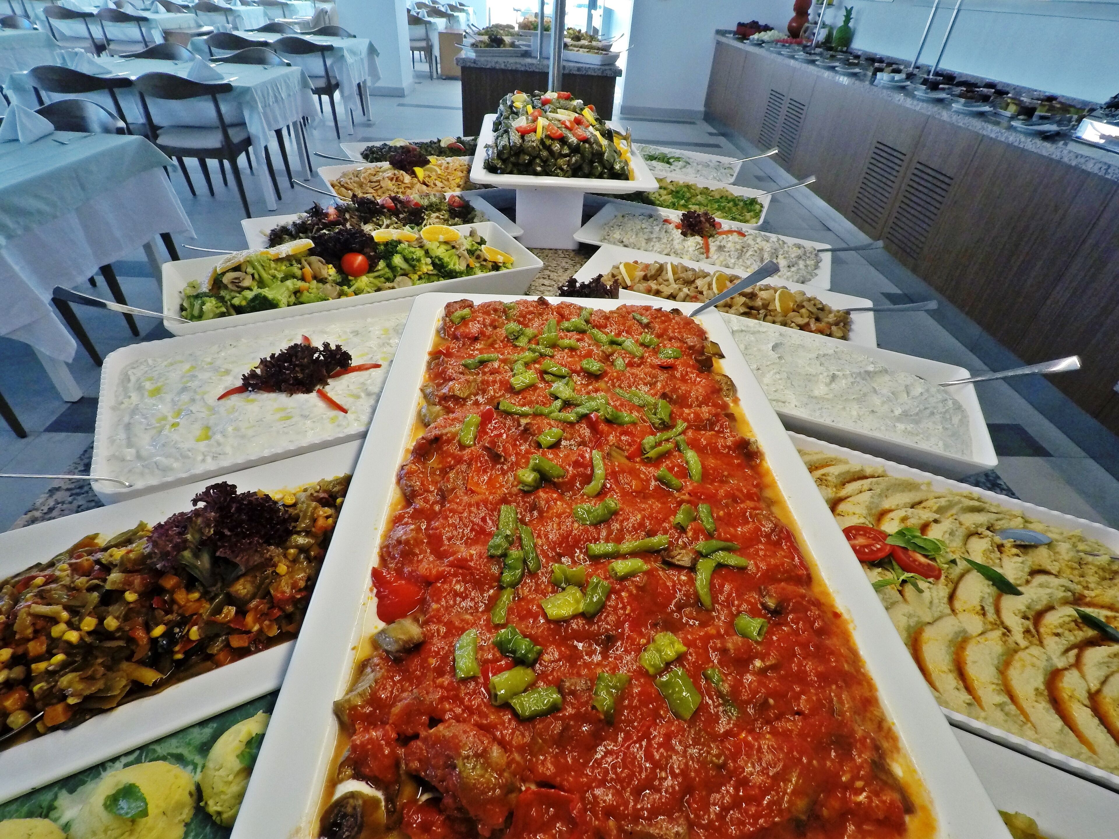 A buffet with various dishes, including pasta, salads, and dips, arranged on tables in a spacious room with chairs.