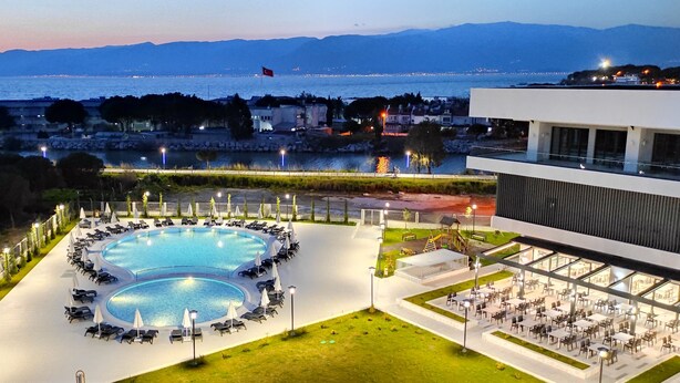 Zona de piscina con sillas de descanso y sombrillas, comedor con mesas y sillas, y vista al mar y las montañas de fondo.