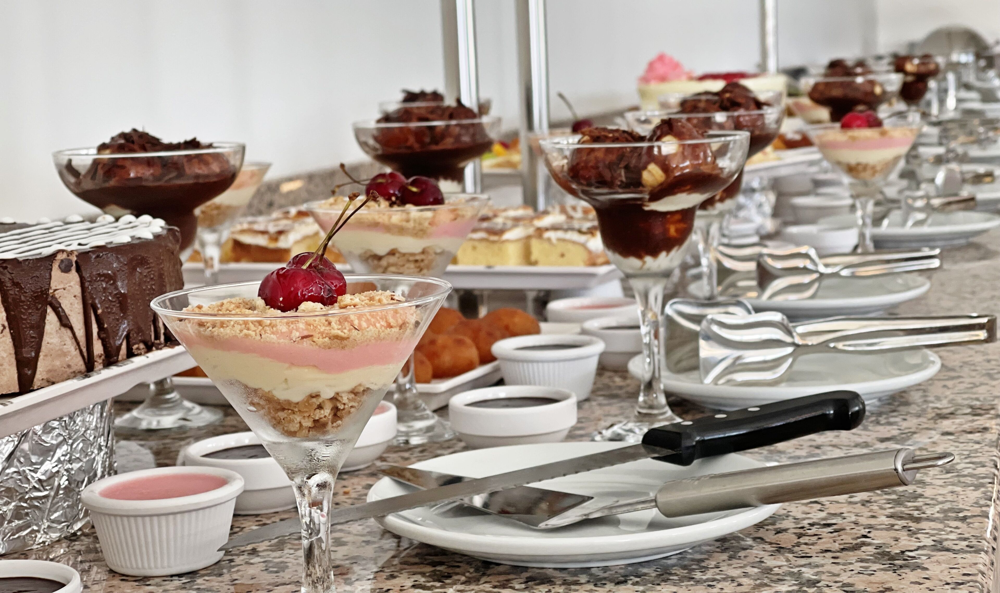 Variedad de postres en una mesa de buffet, incluyendo un pastel de chocolate, un postre en capas en vaso, y varios recipientes pequeños con salsas.