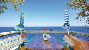 Un banc et une table en bois, recouverts d’un tissu rayé aux couleurs vives, deux parasols rayés et une vue sur la mer.