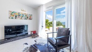 A modern living room with a flat-screen TV, a leather chair, a cowhide rug, and a large window with a view of greenery and a body of water. - Amazing Home in Naklice With Wifi and 2 Bedrooms (Omis)