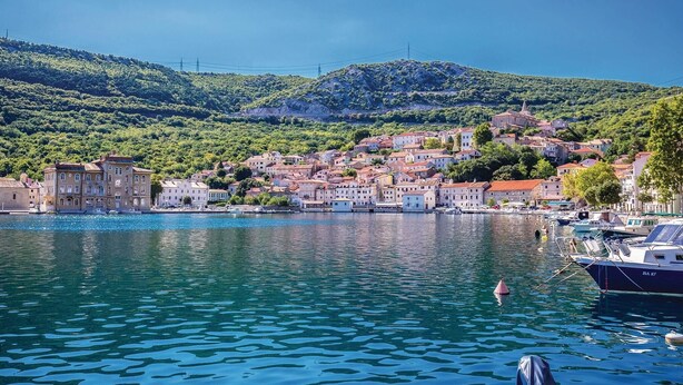 Una cittadina costiera con edifici lungo la passeggiata, acqua cristallina e colline sullo sfondo.