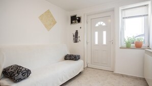 Une pièce blanche avec un canapé blanc, une porte, une fenêtre avec des plantes et un tableau accroché au mur.