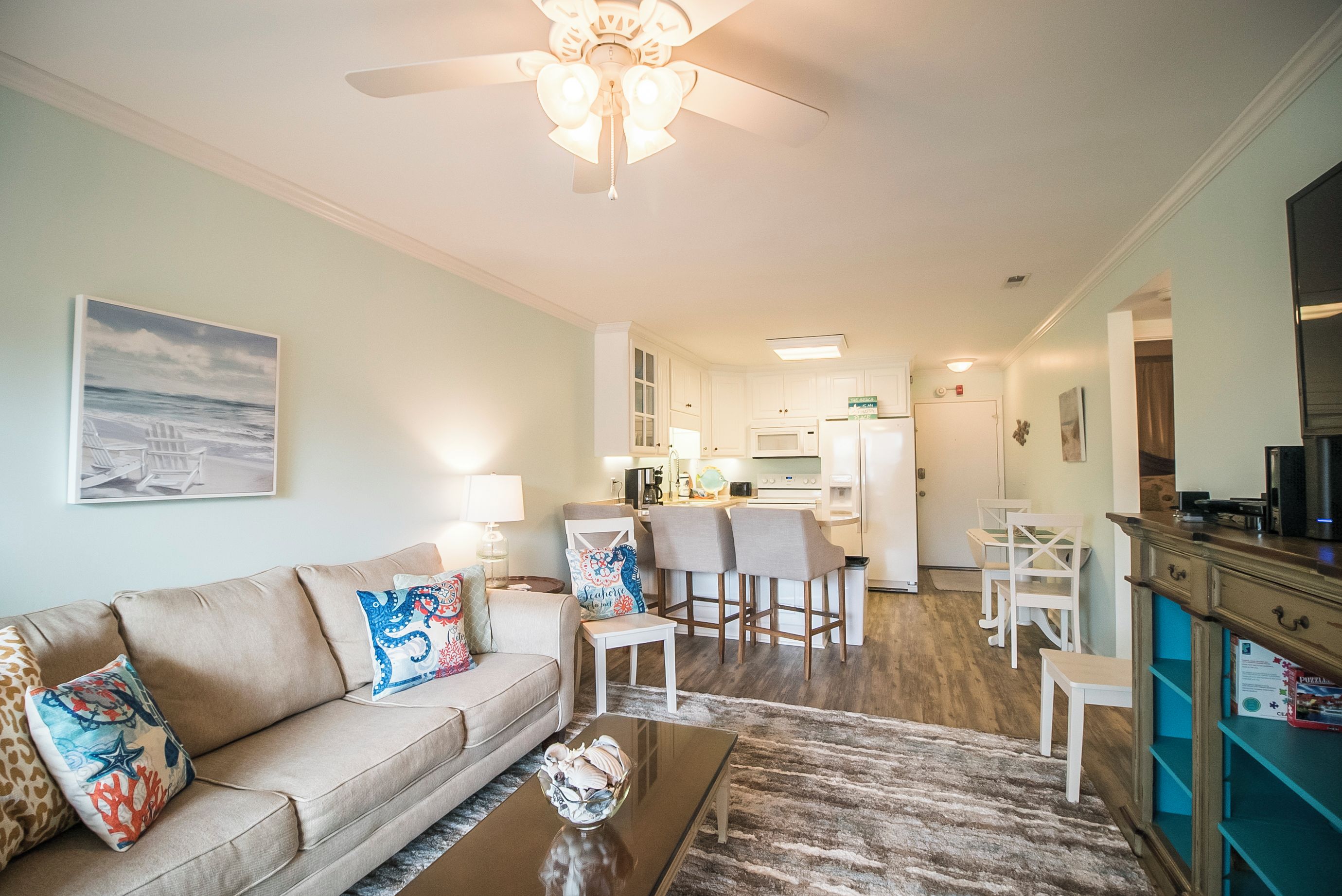 A living room with a sofa, coffee table, and a ceiling fan. There is a kitchen area with a refrigerator and bar stools.