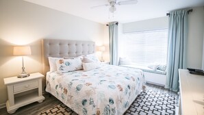 Een slaapkamer met een gestoffeerd hoofdeinde, een bed met een decoratief sprei, een nachtkastje met een lamp, een raam met jaloezieën en een plafondventilator.
