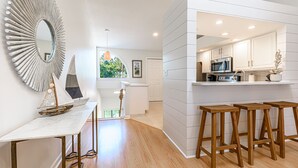 Una cocina moderna con gabinetes blancos, un microondas y un lavamanos. Hay taburetes de madera y un espejo grande en la pared.