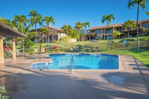 Una piscina con una terraza alrededor, palmeras y un edificio con balcones al fondo.