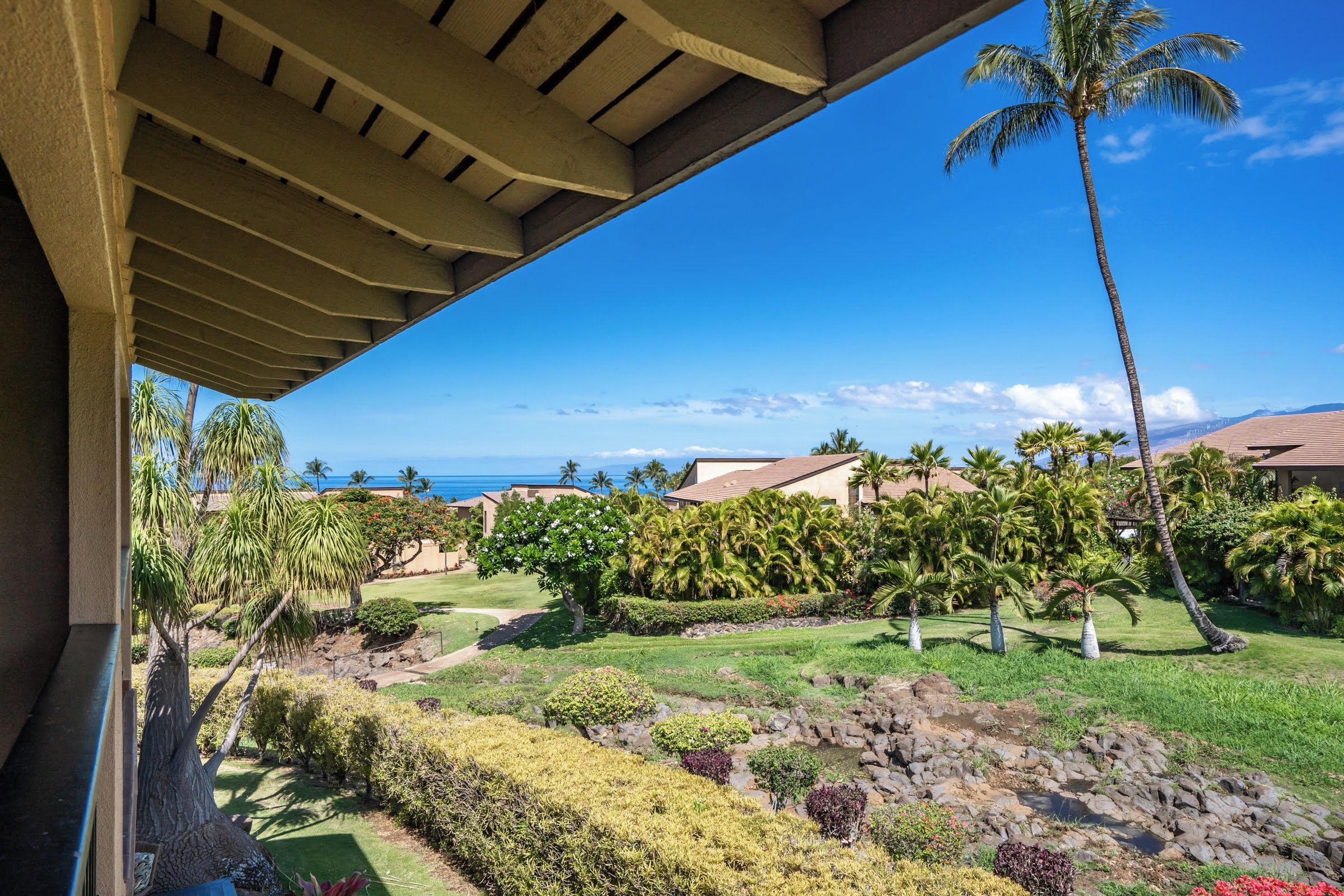 A view from a balcony overlooking a landscaped garden with palm trees, a clear blue sky, and a distant view of the ocean.