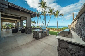 Een terras buiten met een barbecue en zithoek, met uitzicht op een strand met palmbomen en een heldere lucht.