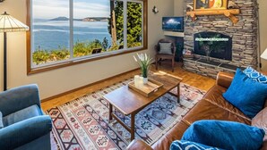A living room with a stone fireplace, a large window with a view of the sea, a patterned rug, and a blue sofa. - Waterfront w/ Seasonal Dock! (Rendezvous Retreat) (Friday Harbor)
