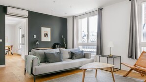 A modern living room with a grey sofa, a small table, and a chair. There is a window with curtains, a radiator, and a picture on the wall. - Modern Landstraße 1BR, walk to Metro U1 and Hauptbahnhof, by Blueground (Vienna)