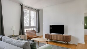 A modern living room with a grey sofa, a wooden coffee table, a wicker chair, a TV on a wooden stand, and a window with curtains. - Modern Landstraße 1BR, walk to Metro U1 and Hauptbahnhof, by Blueground (Vienna)