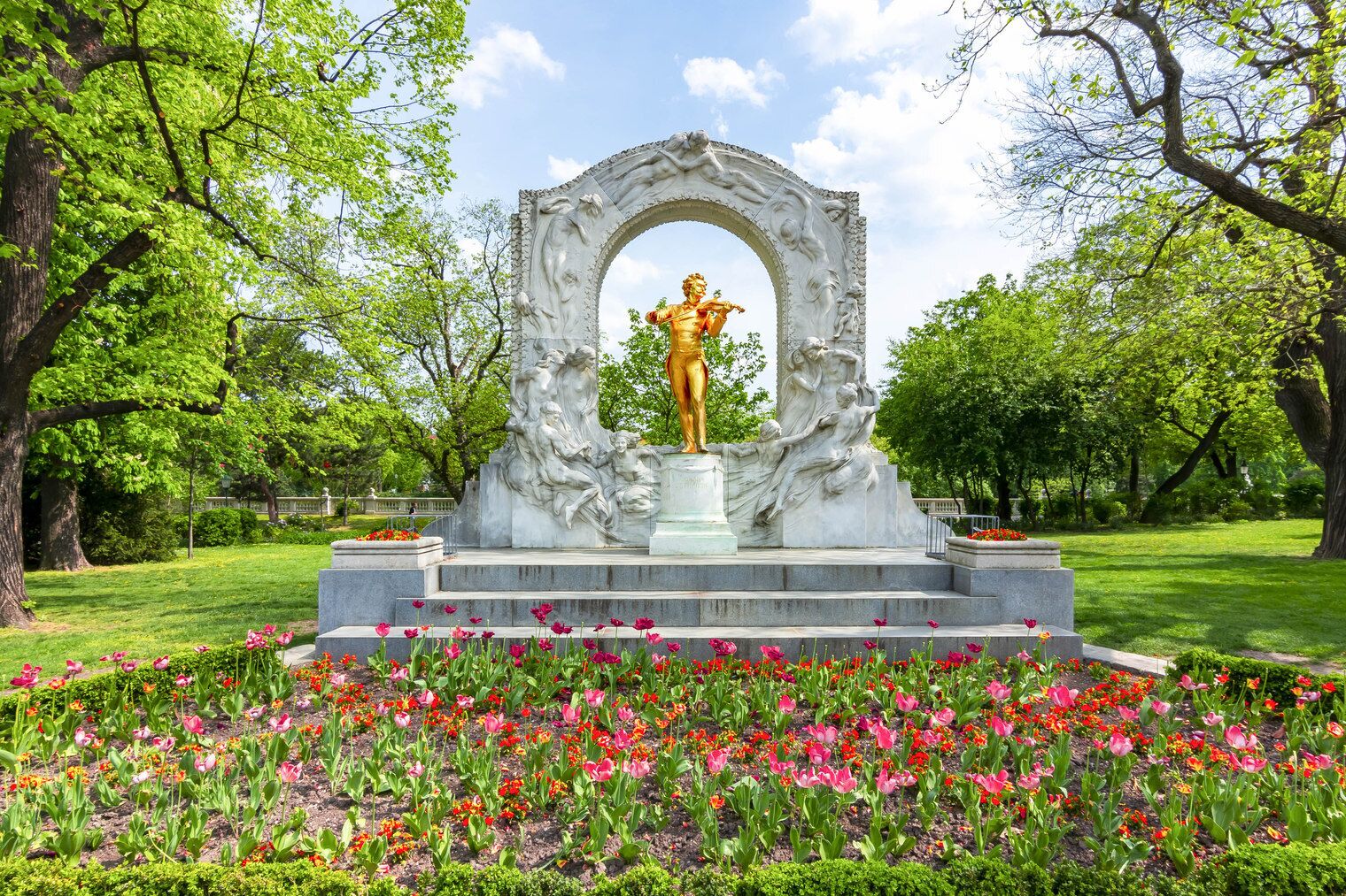 A statue with a golden figure in a white arch, surrounded by a garden with pink and red flowers.