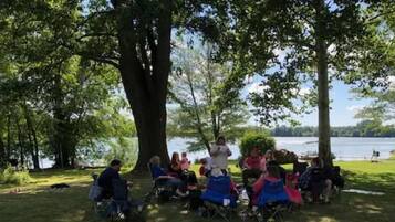 Un grupo de personas sentadas en sillas de jardín bajo árboles grandes junto a un lago.