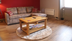 A living room with a wooden coffee table, a sofa with plaid cushions, and a red wall with two framed pictures. - 1 Bed Flat - Sleeps 2 - Wifi (Pitlochry)