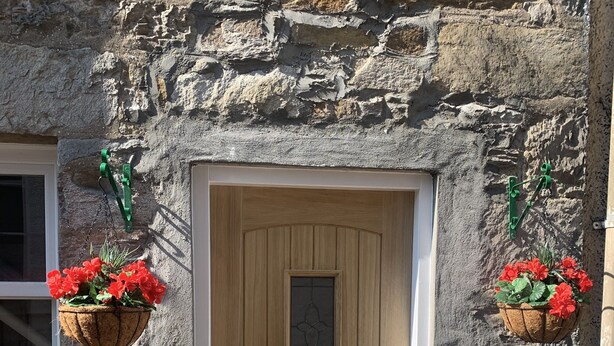 A stone building with a wooden door, two hanging flower baskets, and a window to the left. - 1 Bed Flat - Sleeps 2 - Wifi (Pitlochry)