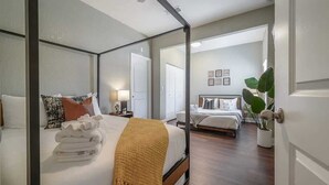 Een hemelbed met een witte deken en een mosterdgele plaid, een nachtkastje met een lamp, en zicht op een aangrenzende slaapkamer met een vergelijkbaar bed.