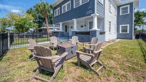 Una casa de dos pisos con un porche cubierto, una fogata y sillas Adirondack.