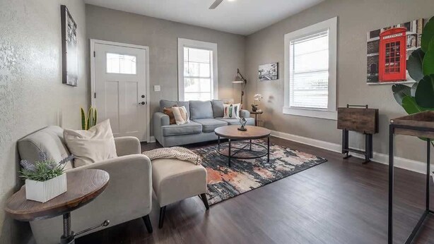 Una sala de estar con un sofá gris, una mesa de centro y una lámpara de pie. Hay dos ventanas y una puerta.