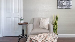 Una habitación con una silla, una mesita, una planta en maceta y una obra de arte colgada en la pared.