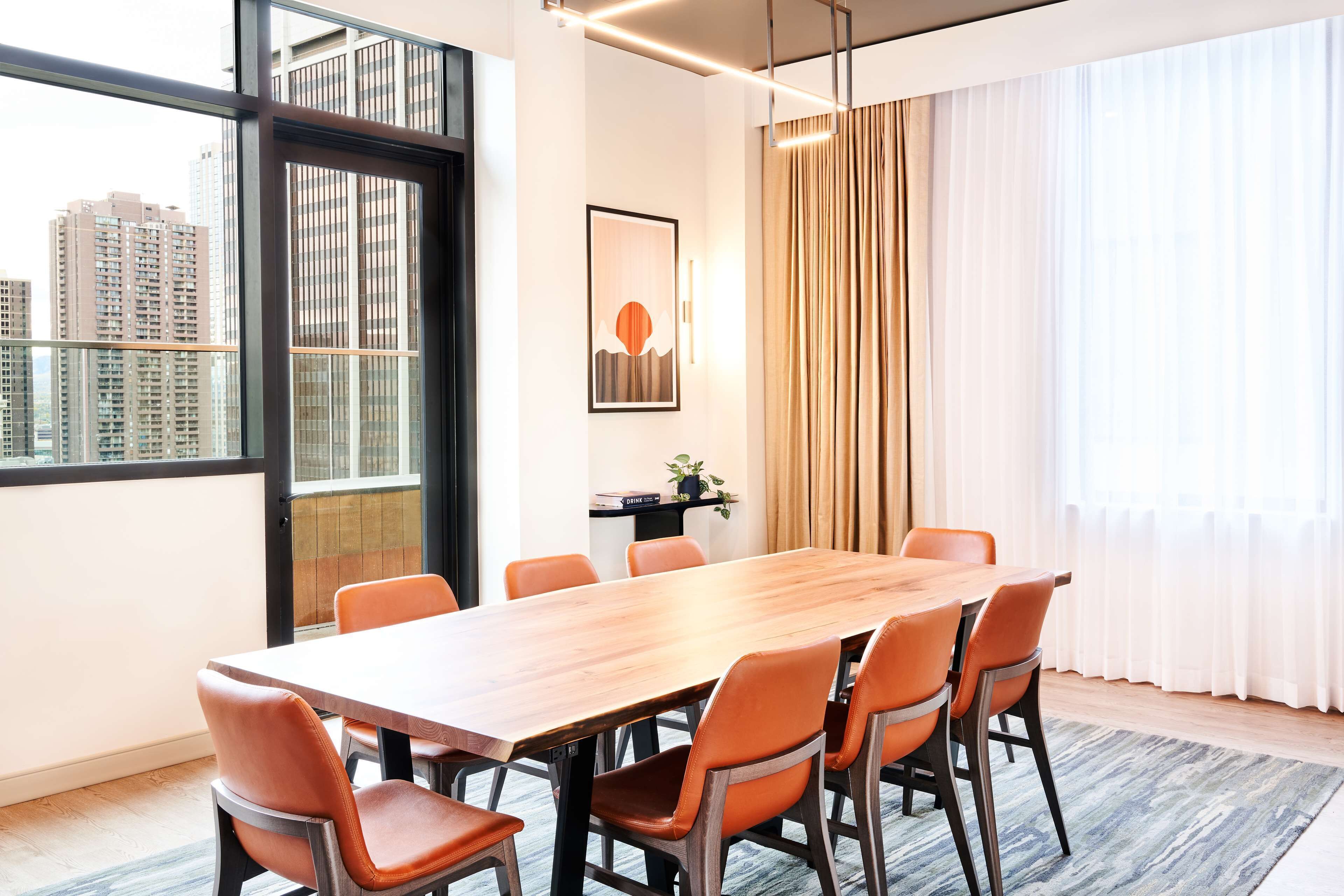 Un salón de conferencias con una larga mesa de madera, seis sillas anaranjadas, un cuadro en la pared y vistas de edificios altos a través de amplias ventanas.