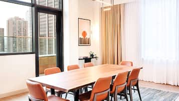A conference room with a long wooden table, six orange chairs, a framed picture on the wall, and a view of tall buildings through large windows.