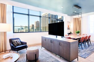 Una sala de estar moderna con un televisor grande, un comedor y vistas de la línea del horizonte de la ciudad a través de amplias ventanas.