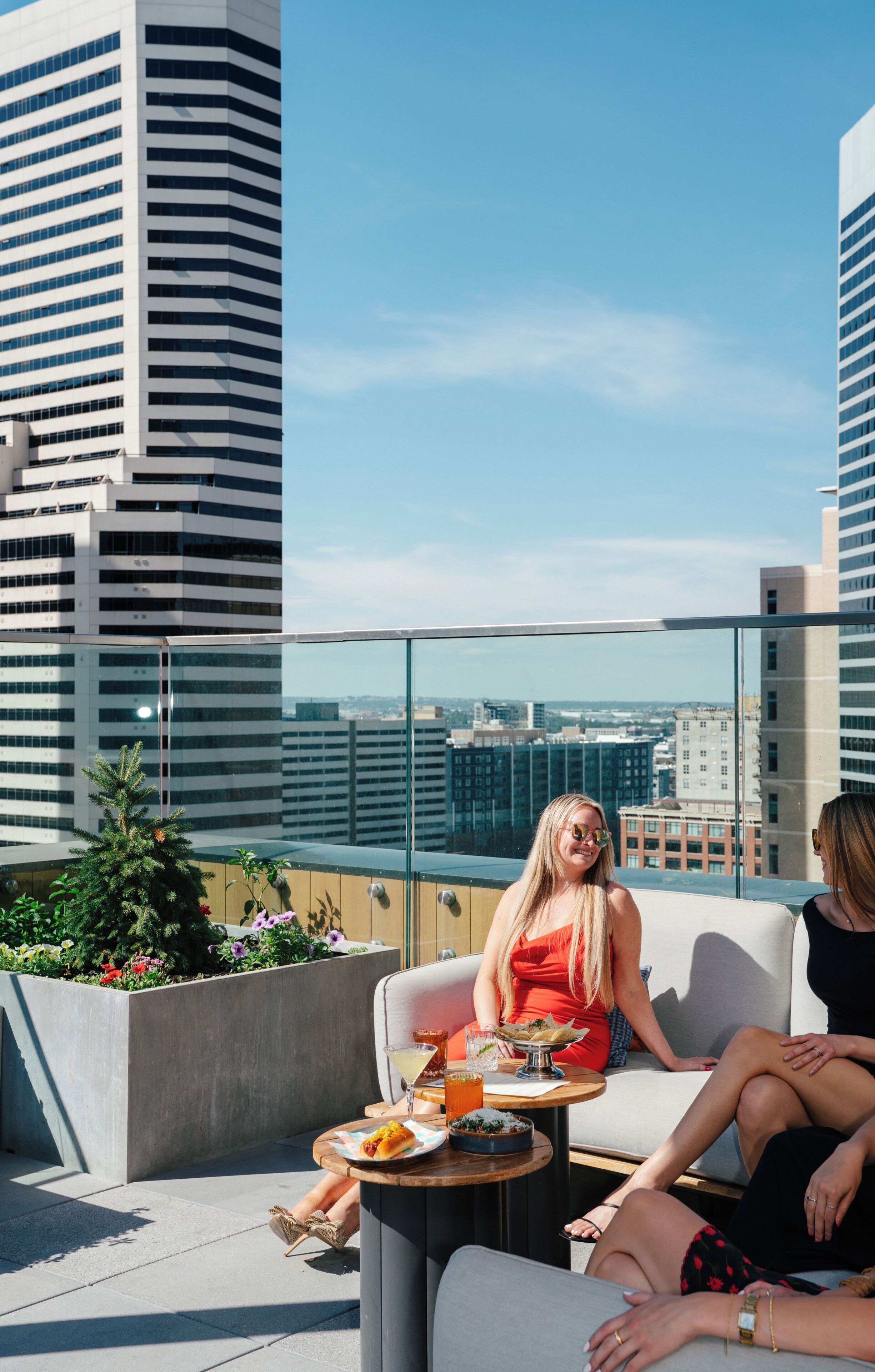 Una terraza en la azotea con barandilla de cristal, una mesa de comedor con una bebida y vistas a la ciudad.