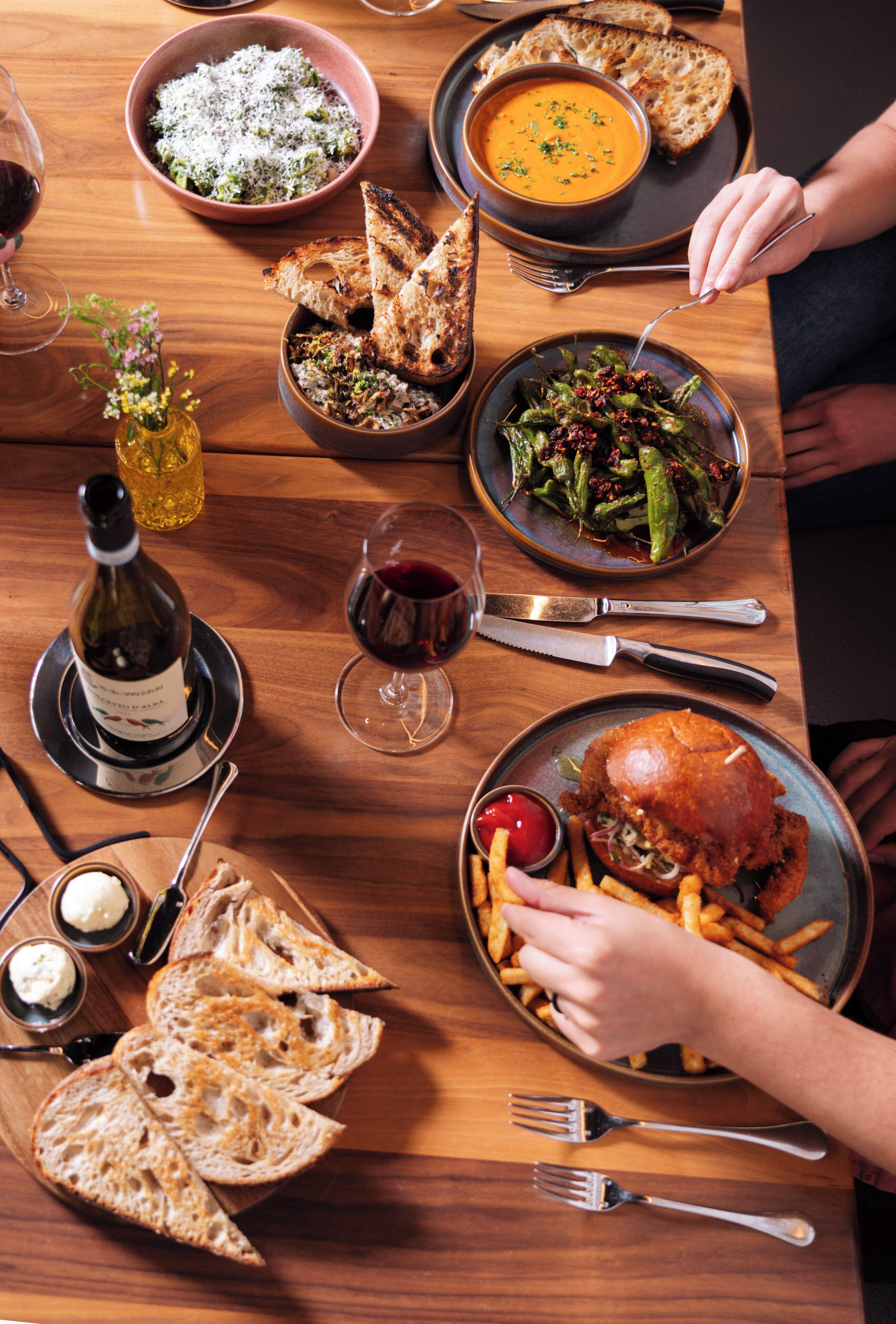 A wooden dining table with various dishes, including a burger, fries, soup, and bread.