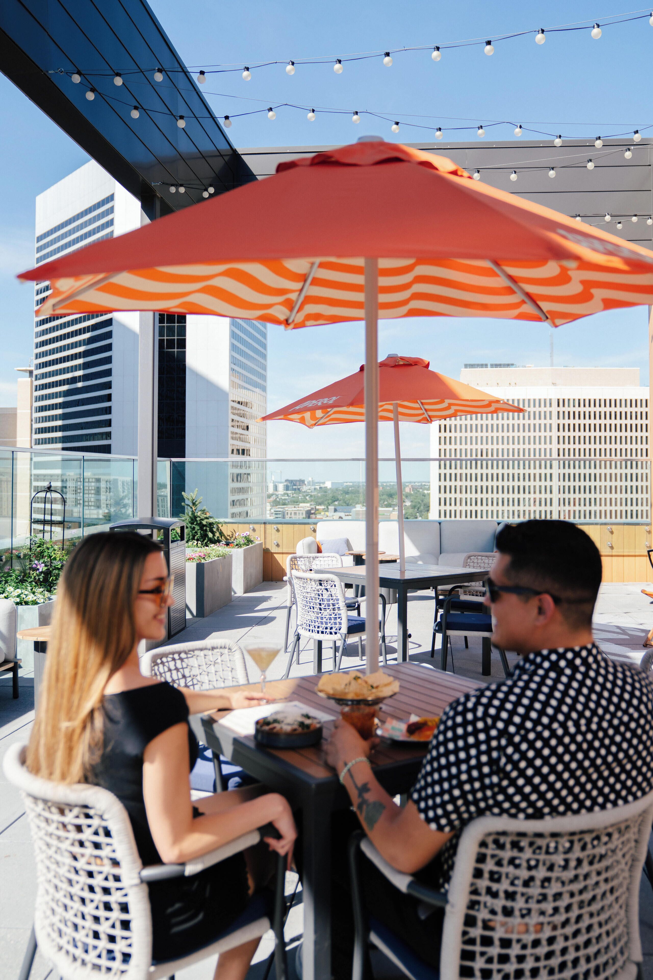An outdoor dining area with tables and chairs, a red umbrella, and a city view.