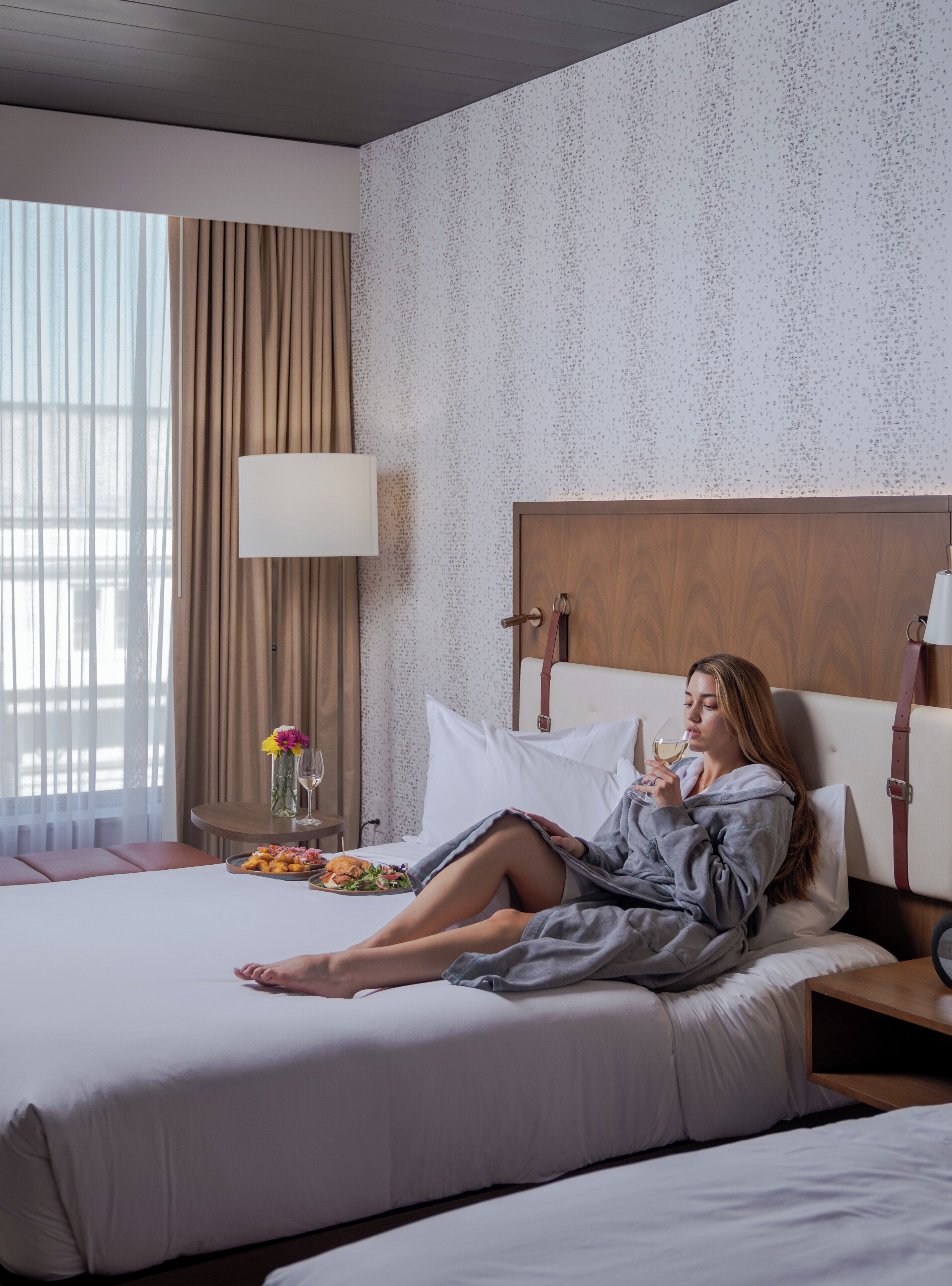 Habitación de hotel con cama, mesita de noche, lámpara, ventana con cortinas y pared con diseño salpicado.