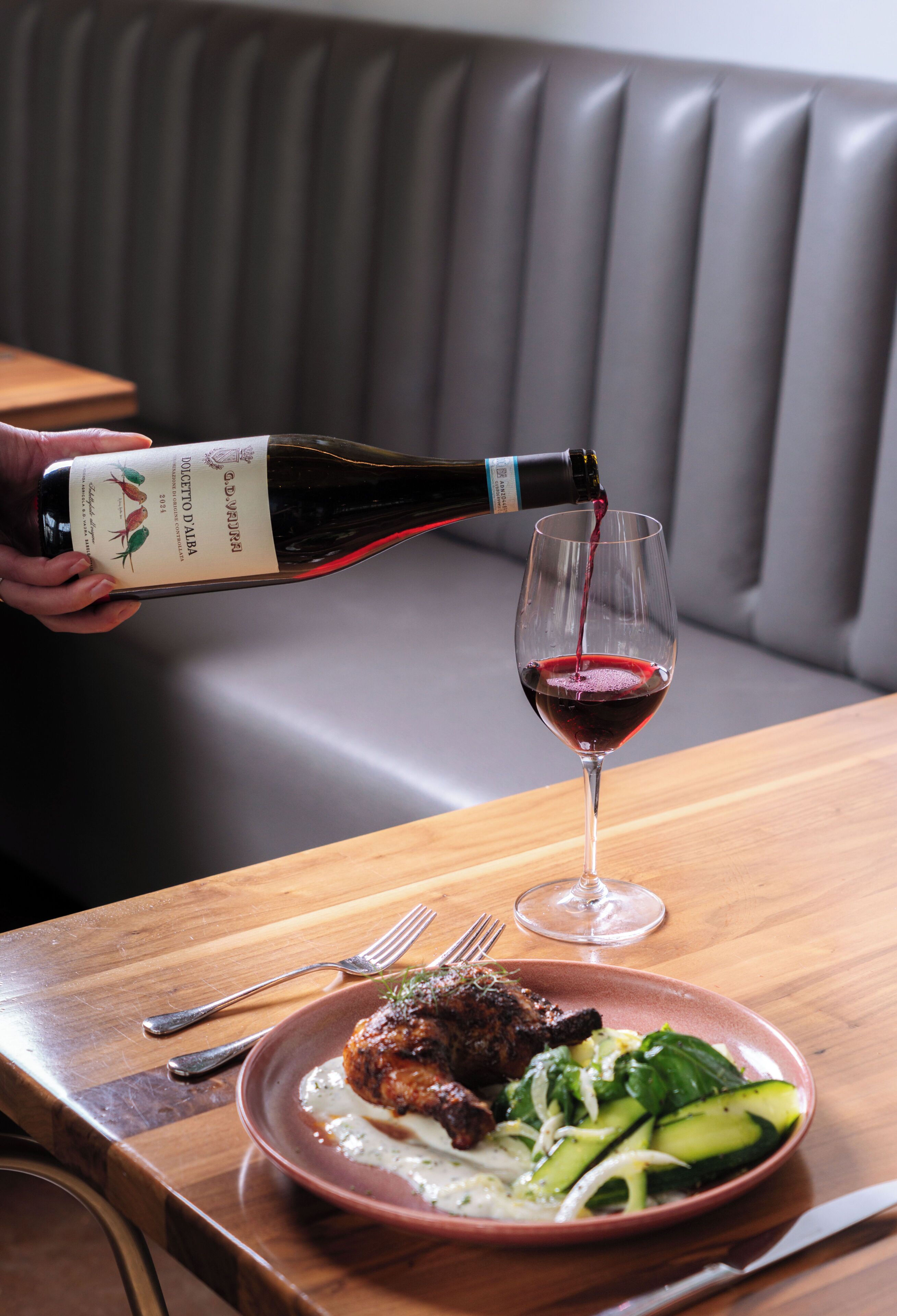 A bottle of wine being poured into a glass on a wooden table with a plate of food.
