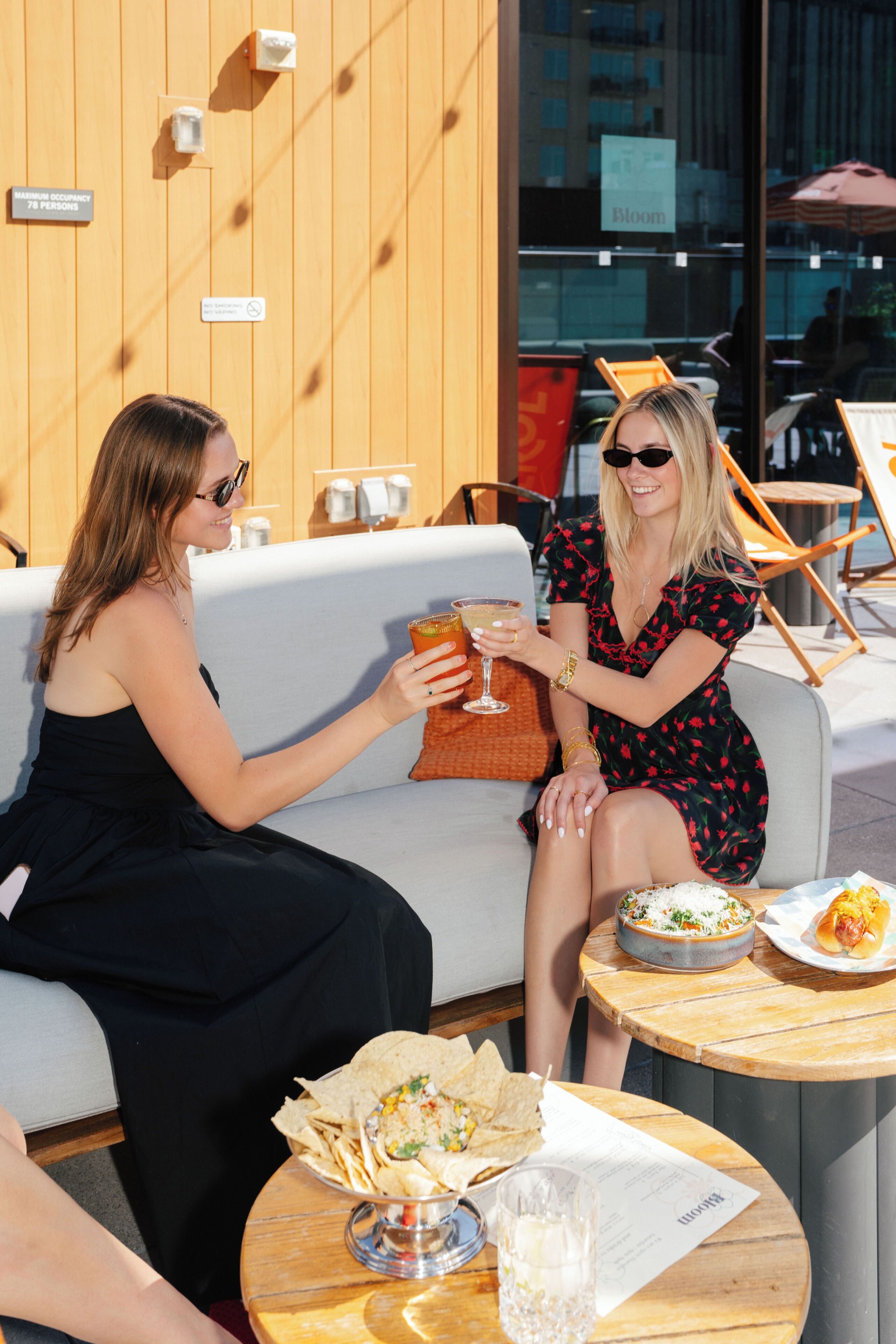 Two people seated at an outdoor table with food and drinks, a wooden wall, and a sign indicating a maximum occupancy of 78 persons.
