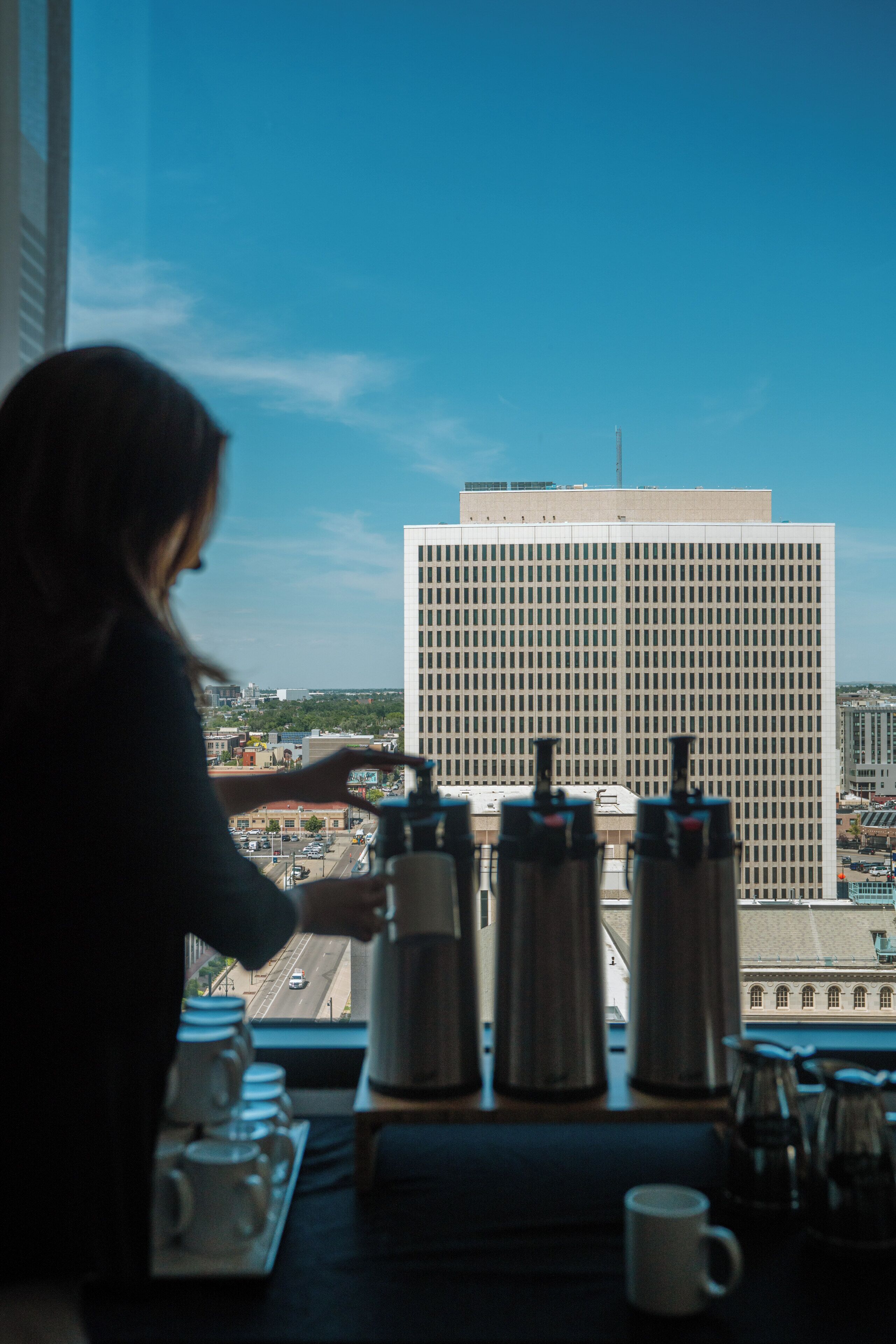 Una persona junto a la ventana con vista a la ciudad, una cafetera y una bandeja con tazas.