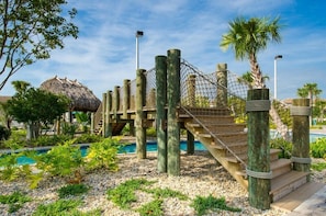 A rope bridge over a pool area with a thatched-roof structure in the background.