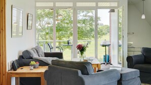 En moderne stue med sofa, spisebord og udsigt til en have gennem store vinduer.