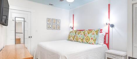 Un dormitorio con cama, cómoda de madera, ventilador de techo y una mesita.